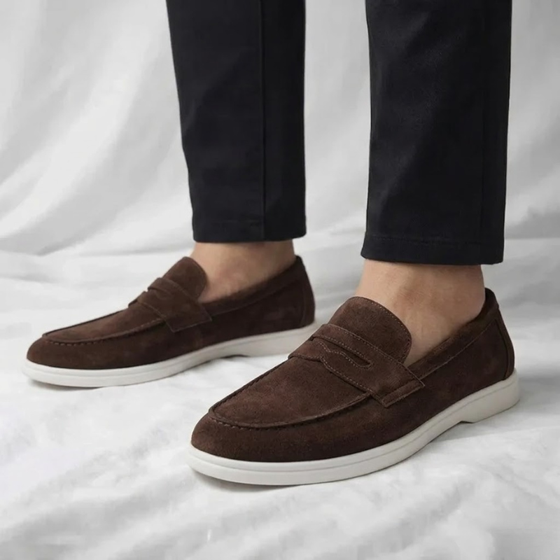 Mocasín de hombre de ante marrón oscuro, con tira cruzada y suela blanca.