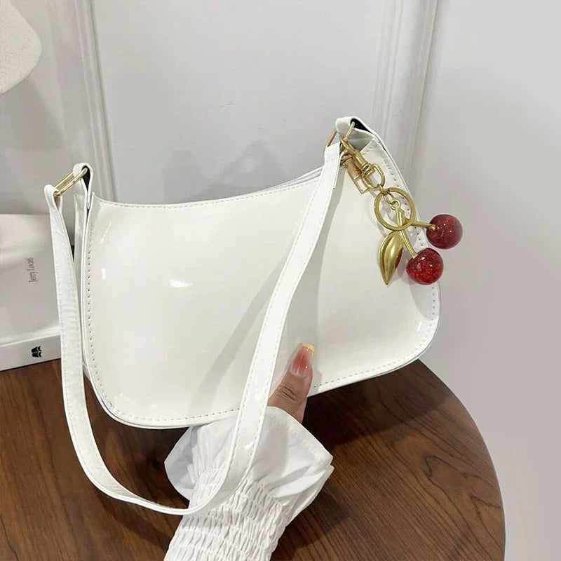 Bolso bandolera de charol blanco con asa larga y detalle de cadena con colgante.