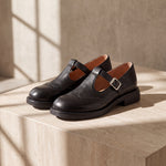 Mocasín Louisa™ de cuero negro con hebilla, par sobre superficie iluminada
